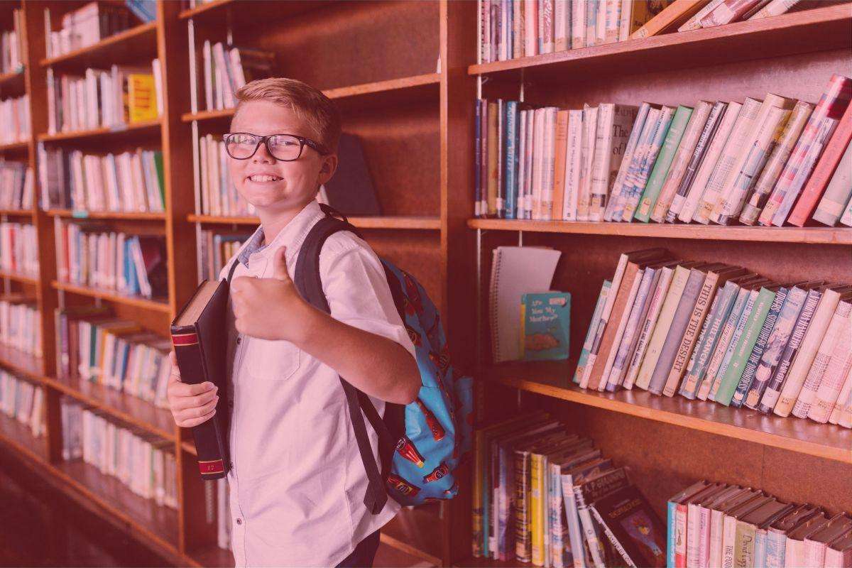 boy standing by a bookshelf with books on, holding a book in one arm and giving a thumbs up sign